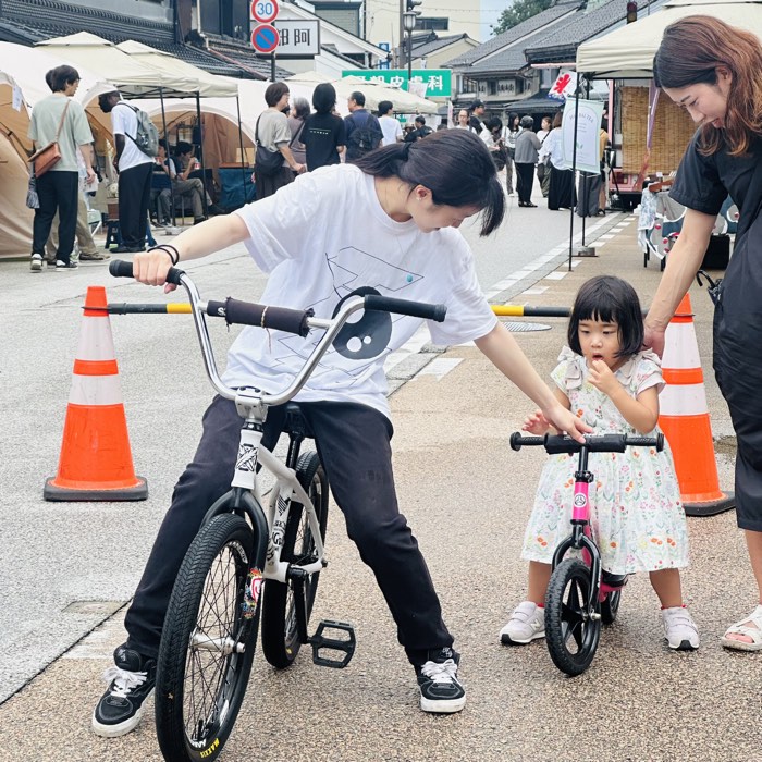 高岡・市場街の体験会でストライダーのハンドルを持つ子どもと、隣でサポートするBMXライダー